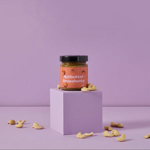 Glas Nutbutter Bruschetta von ae kitchen auf lila Podest, umgeben von Cashews, vor lila Hintergrund