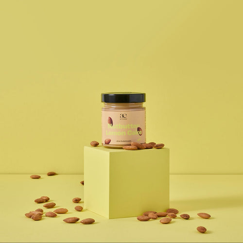 Glas Nutbutter Lemon Cake von ae kitchen auf gelbem Podest, umgeben von Mandeln, vor gelbem Hintergrund