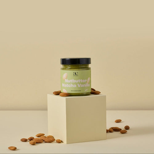 Glas Nutbutter Matcha Vanilla von ae kitchen auf cremefarbenem Podest, umgeben von Mandeln, vor beigem Hintergrund