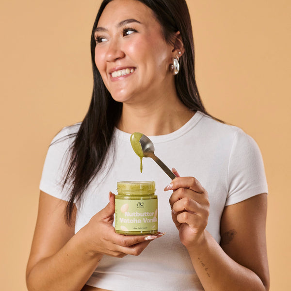 Frau hält lachend ein offenes Glas Nutbutter Matcha Vanilla von ae kitchen und zieht einen tropfenden Löffel heraus