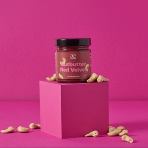 Glas Nutbutter Red Velvet von ae kitchen auf pinkem Podest, umgeben von Cashews, vor pinkem Hintergrund
