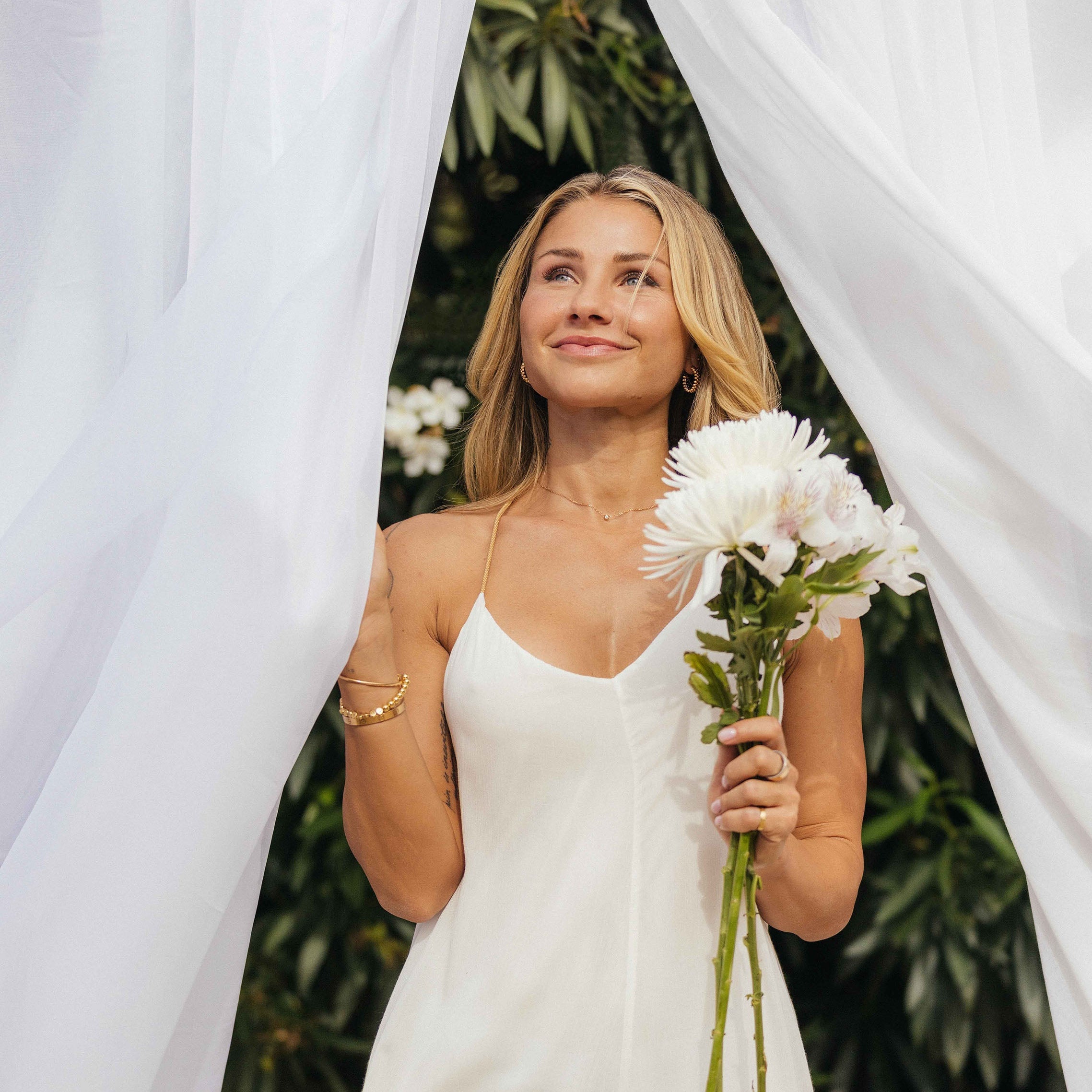Eine Frau in einem weißen Kleid steht zwischen wehenden weißen Stoffbahnen und hält einen Strauß heller Blumen. Sie lächelt sanft nach oben, während grünes Blattwerk den Hintergrund bildet.