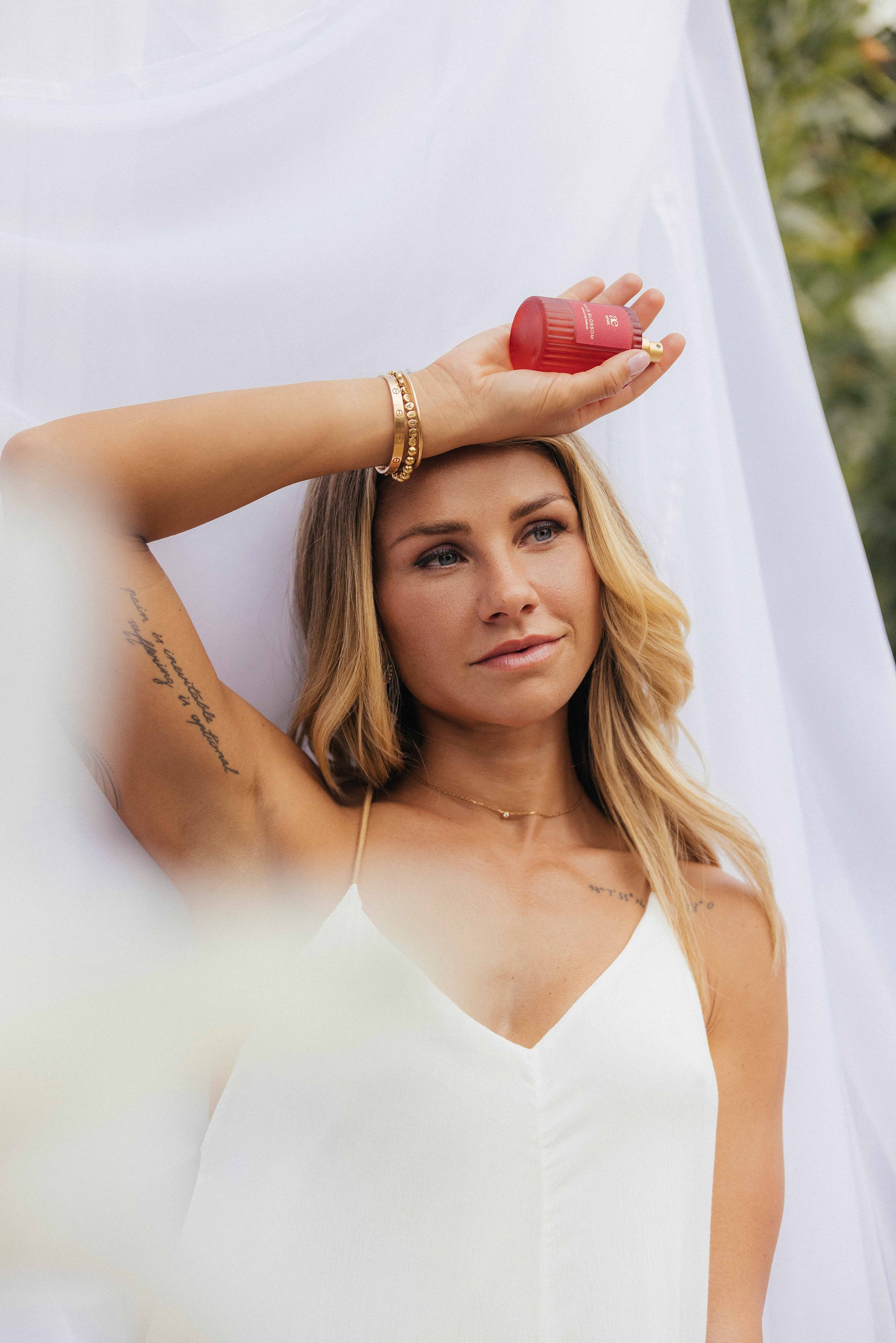 Eine Frau in einem weißen Kleid hält einen kleinen roten Parfumflakon auf ihrem Arm, während weicher weißer Stoff im Vordergrund verschwimmt. Sie blickt ruhig nach vorn, umgeben von einem sanften, hellen Hintergrund.