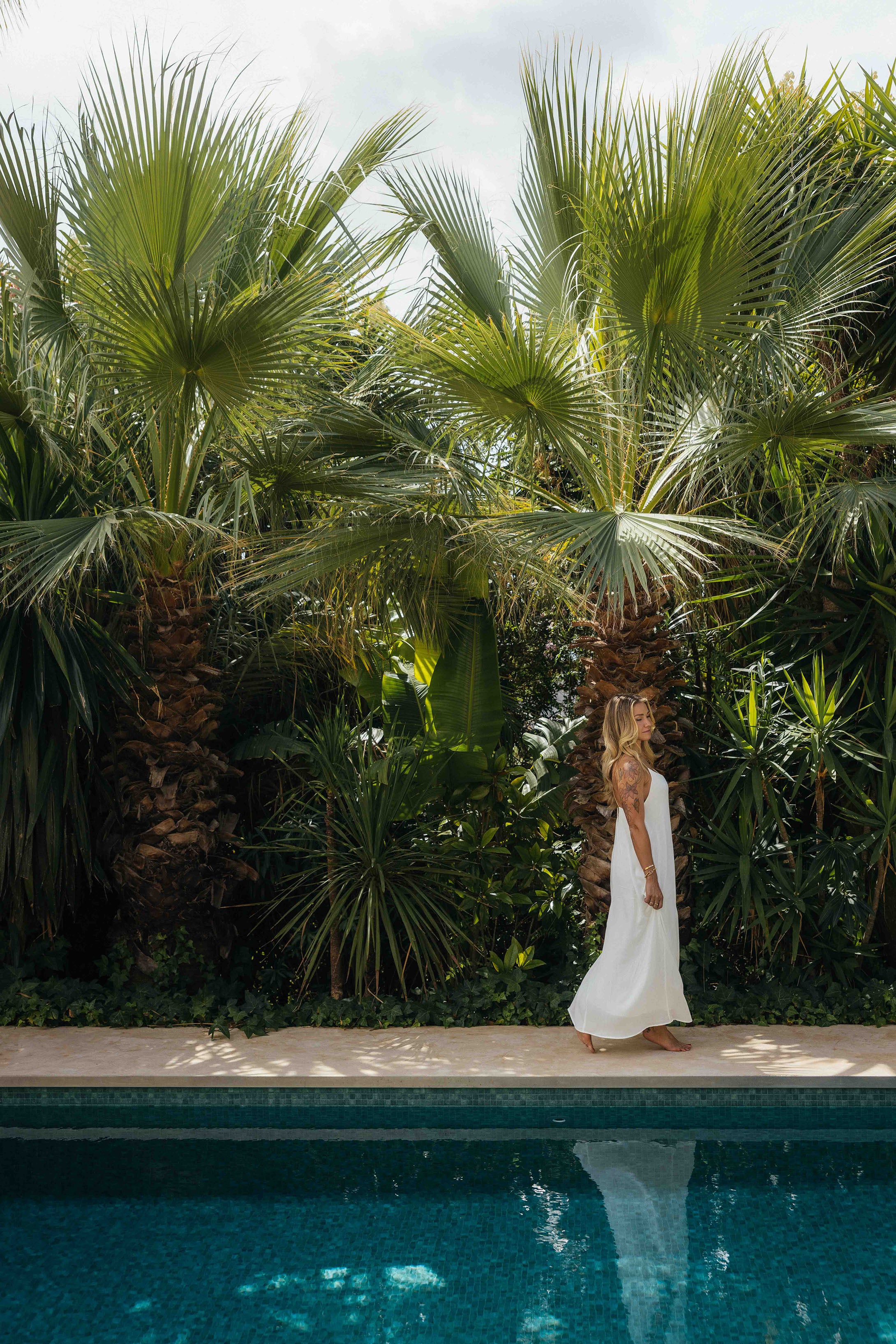 Eine Frau in einem langen weißen Kleid geht barfuß am Rand eines Pools entlang. Hinter ihr steht eine dichte Wand aus großen, tropischen Palmen und üppigem Grün, das sich im Wasser spiegelt.