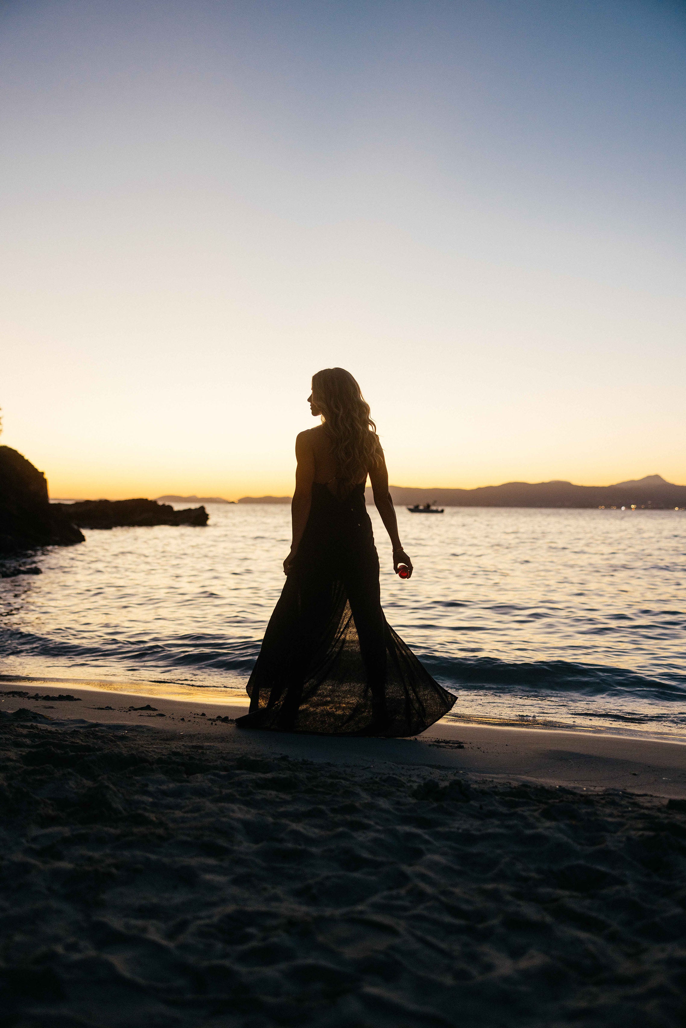 Eine Frau im schwarzen Kleid geht bei Sonnenuntergang am Strand entlang. Ihr Silhouette hebt sich vor dem goldenen Himmel ab, während sanfte Wellen ans Ufer rollen und ein kleiner roter Flakon in ihrer Hand leuchtet.