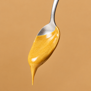 Nahaufnahme eines silbernen Löffels mit cremiger, goldgelber ae KITCHEN Honey Cone Nutbutter, die langsam vom Löffel tropft, vor einem warmen, beigefarbenen Hintergrund.
