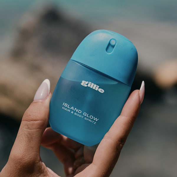Blauer gillie Island Glow Hair & Body Spritz in einer Hand vor sommerlicher Outdoor Kulisse.