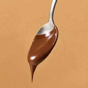 Nahaufnahme eines silbernen Löffels mit cremiger, glänzender Jaffa Cake Nutbutter von ae KITCHEN, die langsam vom Löffel tropft, vor einem warmen, beigebraunen Hintergrund.
