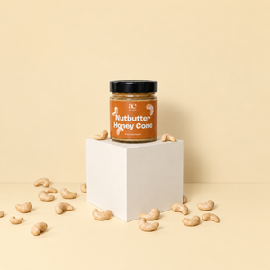 Ein Glas ae KITCHEN Nutbutter Honey Cone mit braunem Etikett und schwarzem Deckel steht auf einem cremefarbenen Podest. Rundherum liegen Cashewkerne vor einem hellen, beigen Hintergrund.