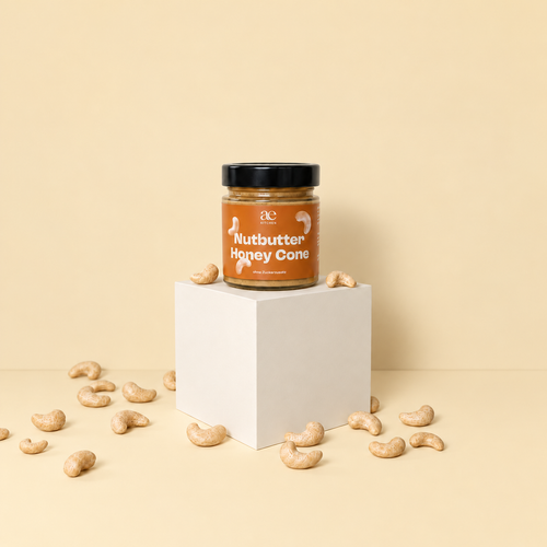 Ein Glas ae KITCHEN Nutbutter Honey Cone mit braunem Etikett und schwarzem Deckel steht auf einem cremefarbenen Podest. Rundherum liegen Cashewkerne vor einem hellen, beigen Hintergrund.