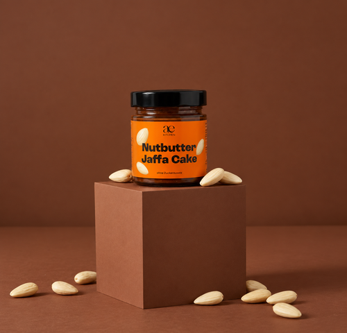 Ein Glas ae KITCHEN Nutbutter Jaffa Cake mit orangefarbenem Etikett und schwarzem Deckel steht auf einem braunen Podest. Rundherum liegen geschälte Mandeln vor einem warmen, braunen Hintergrund.