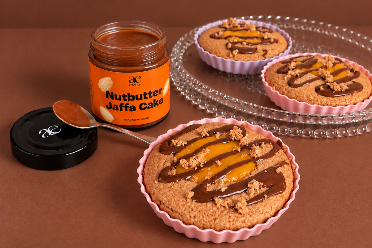Offenes Glas ae KITCHEN Nutbutter Jaffa Cake mit orangefarbenem Etikett, daneben ein Löffel mit cremigem Nussmus und drei kleine Törtchen mit Orangenfüllung, Schokoladendrizzle und knusprigen Streuseln auf warmem, braunem Hintergrund.