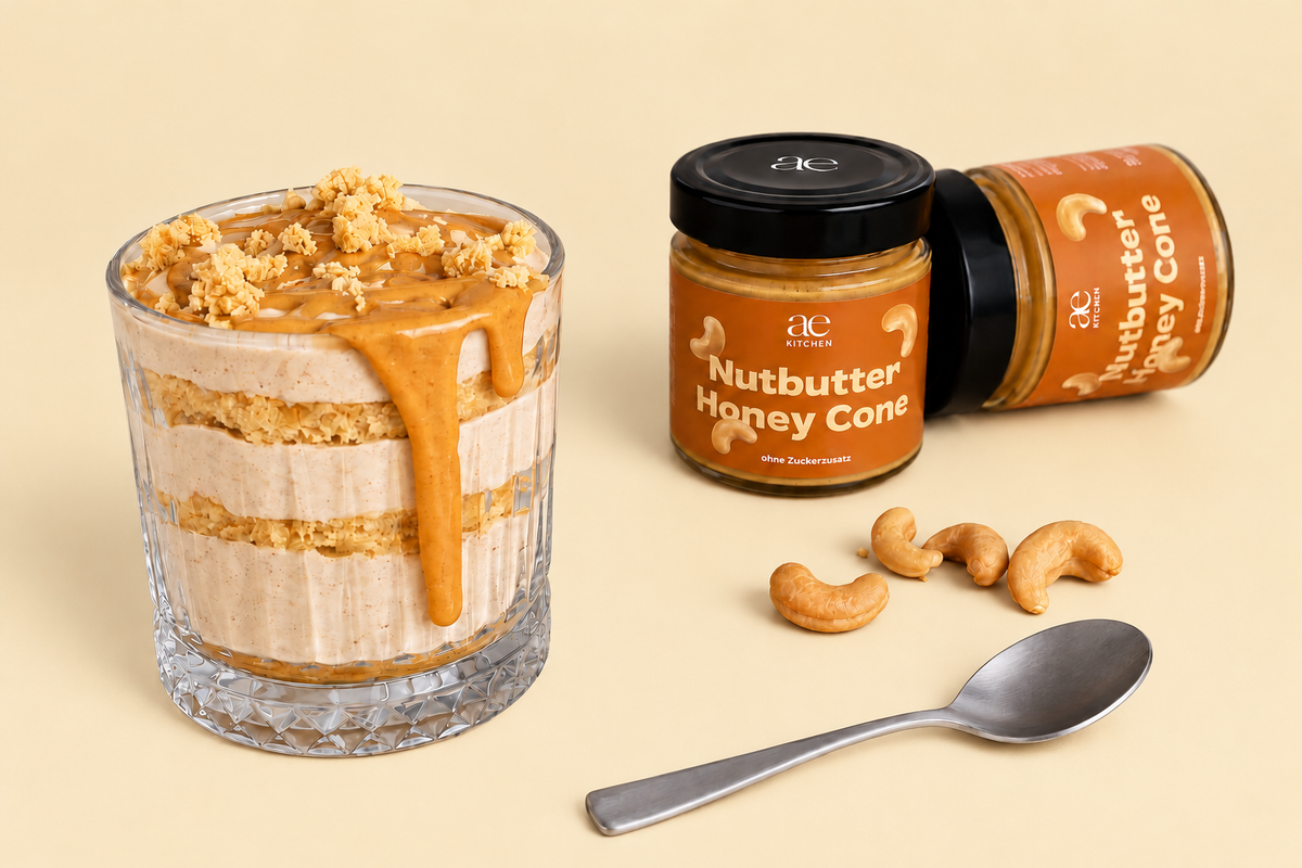 Cremiges Schichtdessert im Glas mit knusprigen Waffelstückchen und ae KITCHEN Honey Cone Nutbutter als Topping. Daneben stehen zwei Gläser Honey Cone Nussmus mit Cashewkernen und einem silbernen Löffel auf beigefarbenem Hintergrund.