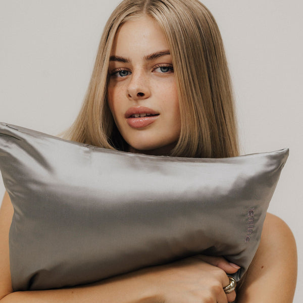 Blonde Frau umarmt unser graues Maulbeerseidenkissen mit gillie-Stickerei und lächelt sanft in die Kamera.